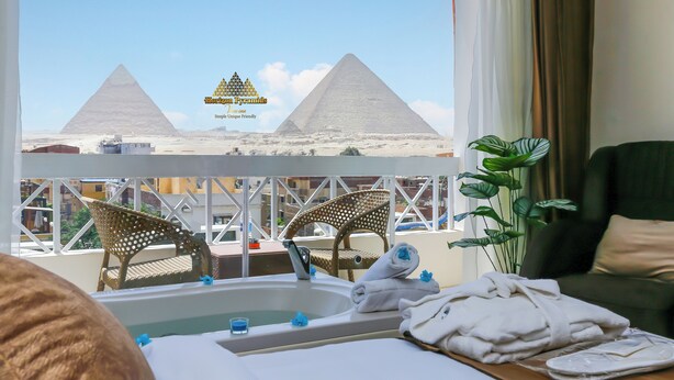 Une chambre d’hôtel avec vue sur les pyramides, une baignoire, deux fauteuils en osier et une plante en pot.