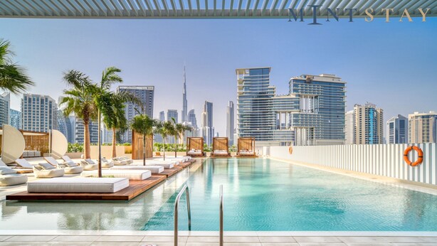 A rooftop infinity pool with lounge chairs and palm trees, overlooking a city skyline with modern buildings. - Charming 1BR w Balcony in Business Bay (Dubai)