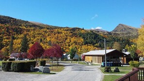 Eine ländliche Straße führt zu einem Gebäude mit einem geneigten Dach, umgeben von herbstlich gefärbten Bäumen und einem Berg im Hintergrund.