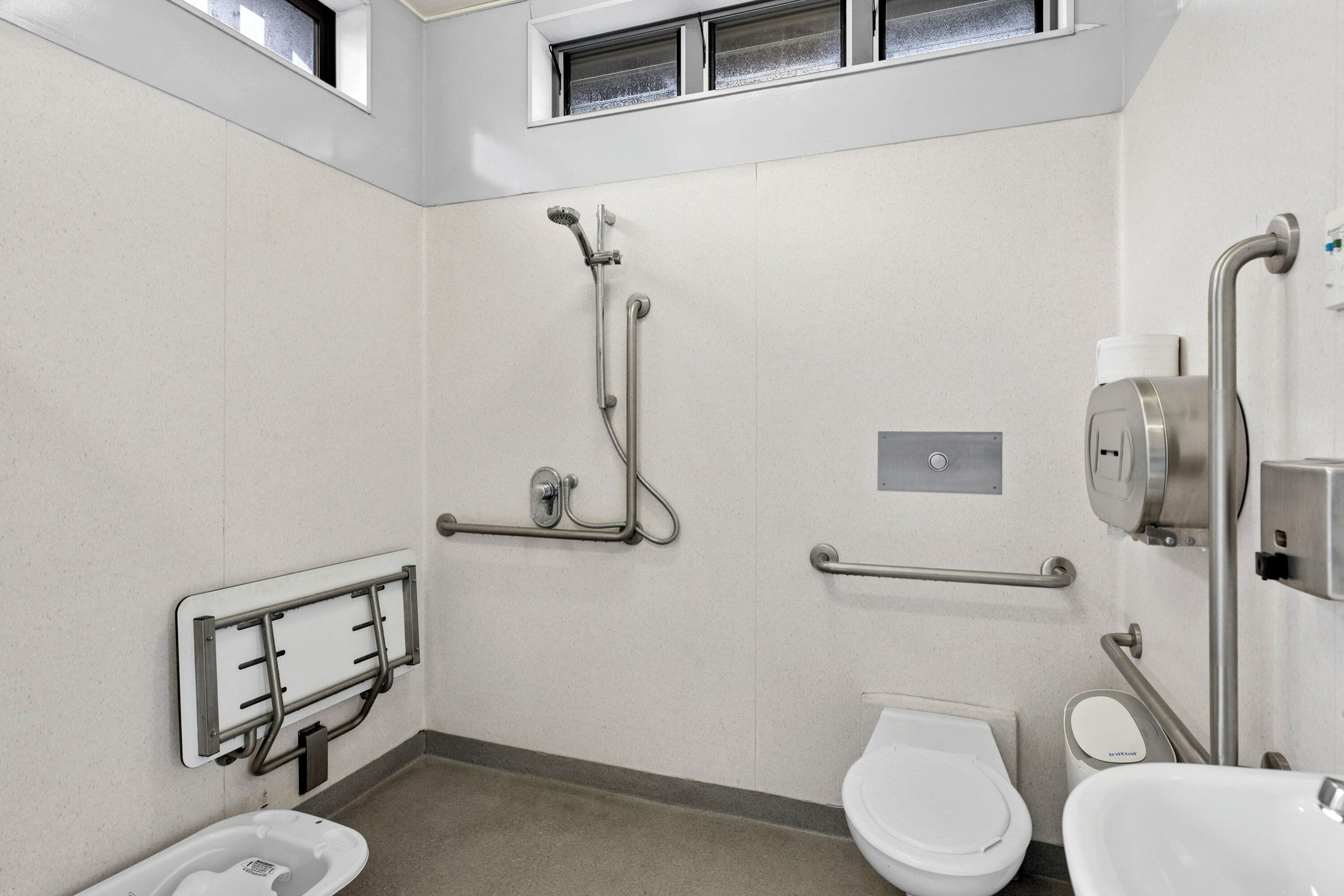 Uma casa de banho moderna com sanita, bidé e chuveiro. Inclui barras de apoio e um cadeiral de chuveiro acessível a cadeiras de rodas.