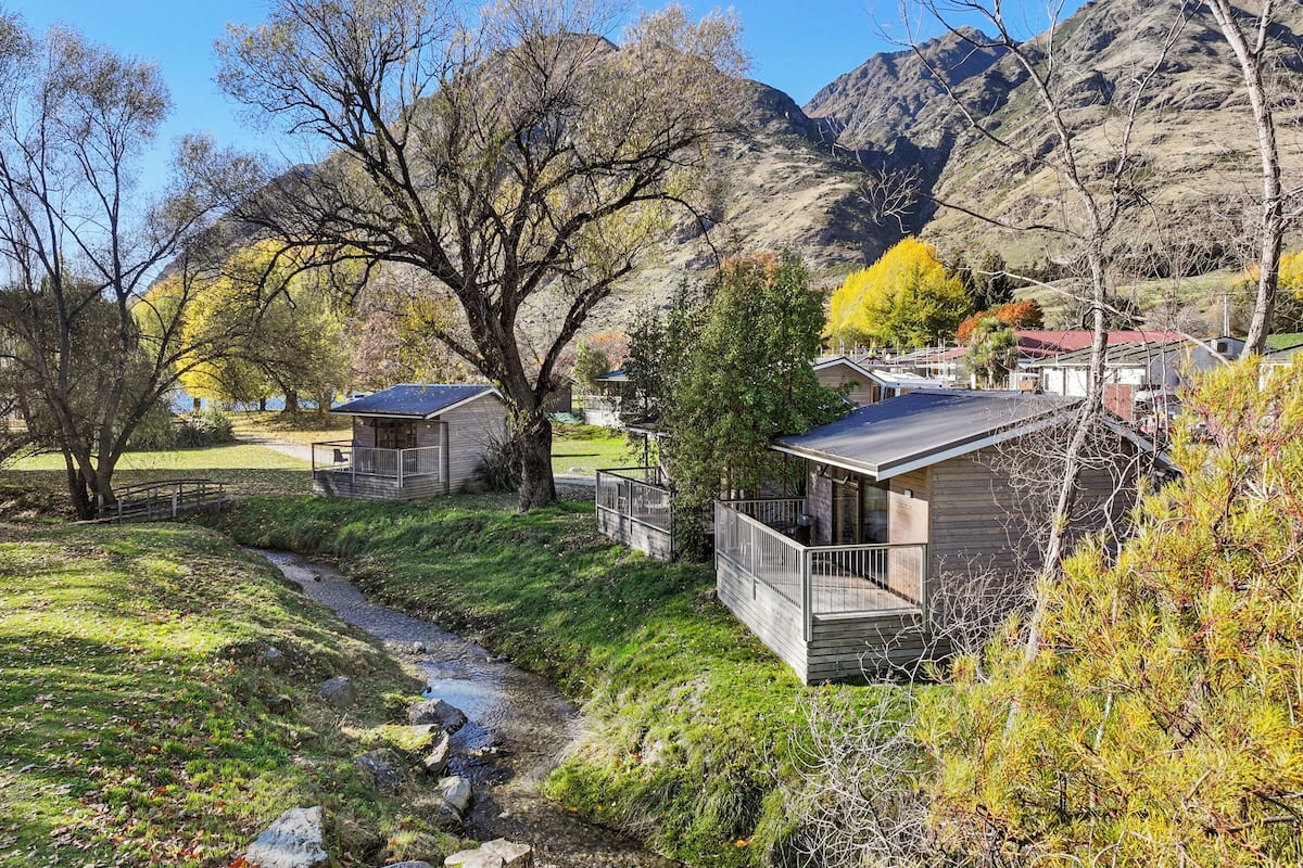 Une cabane dotée d’une terrasse et d’un petit porche, entourée d’arbres et bordée par un ruisseau, avec des montagnes en toile de fond.