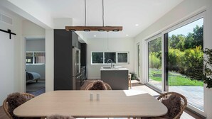 A modern kitchen with a central island, a dining area with a wooden table and wicker chairs, and large sliding glass doors leading to a garden. - Creekside 2-bedroom cottage mid-valley (Hailey)