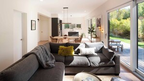 A modern living room with a grey sectional sofa, a dining area with a wooden table and chairs, and a large sliding glass door leading to a patio. - Creekside 2-bedroom cottage mid-valley (Hailey)