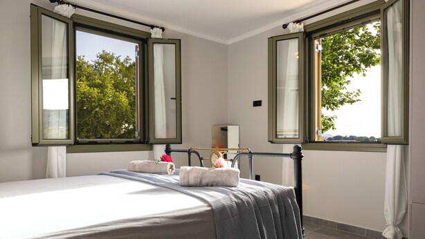 Una cama bien hecha con una manta gris, una mesita de noche con lámpara y dos ventanas abiertas con cortinas.