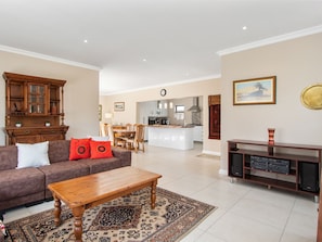 A living room with a sofa, coffee table, and patterned rug. A kitchen with white cabinets and a dining area with chairs is visible in the background. - Martilda Langebaan Country Estate (Langebaan)