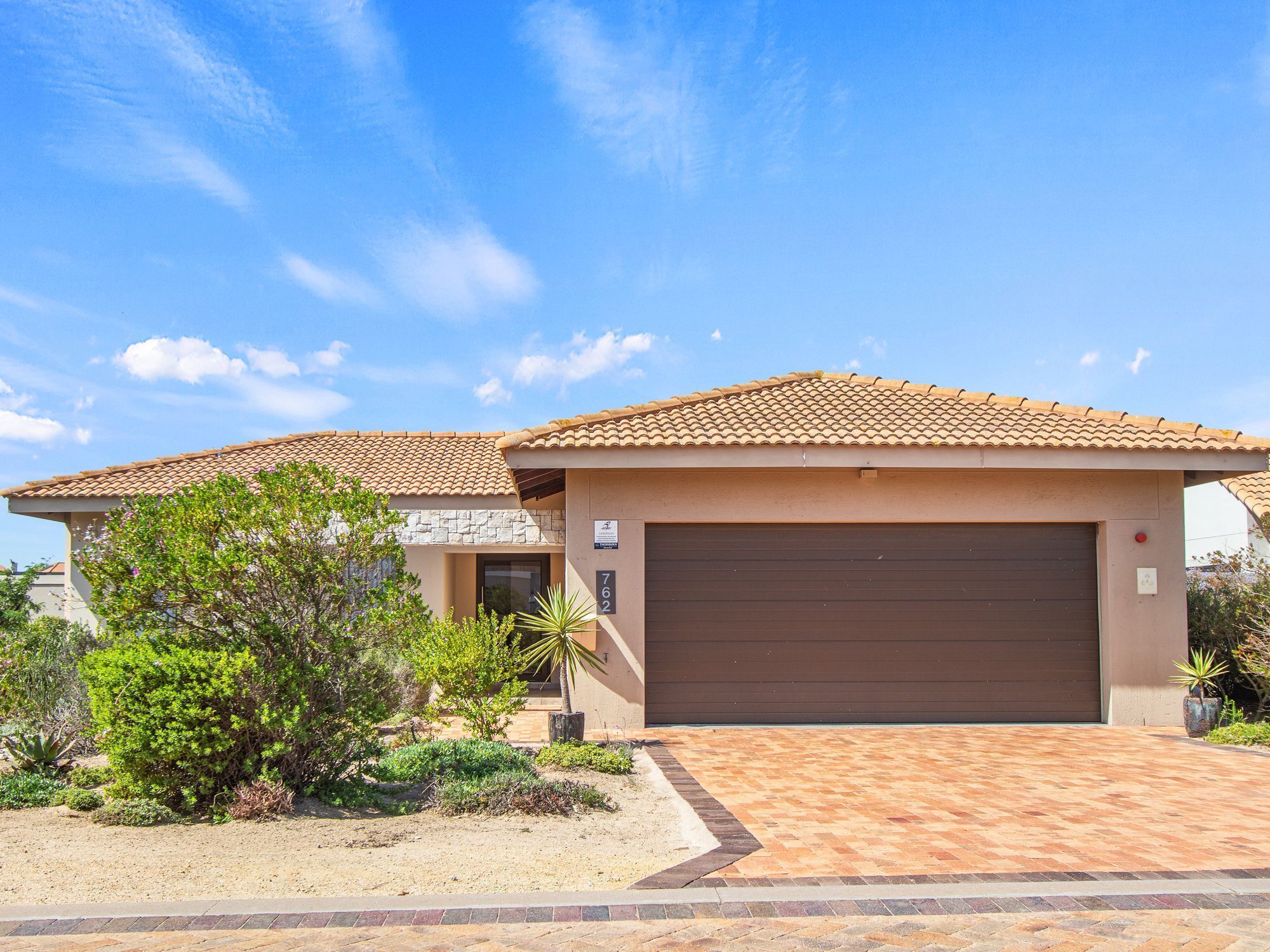 A two-story house with a brown garage door, surrounded by landscaped gardens and a paved driveway.