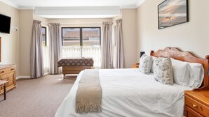 Een slaapkamer met een groot bed, een houten nachtkastje, een televisie, een raam met gordijnen, en een schilderij aan de muur.