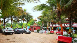 A paved parking area with cars, palm trees, and a small building with a red roof. - We Hotels Alibaug (Alibag)
