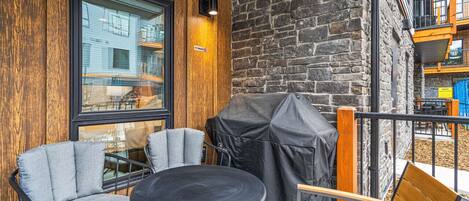 An outdoor seating area with a round table and chairs, a covered grill, and a view of a building through a window.