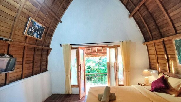 Een slaapkamer met houten plafond, een bed met opgerolde handdoek, een televisie aan de muur en een balkon met gordijnen.