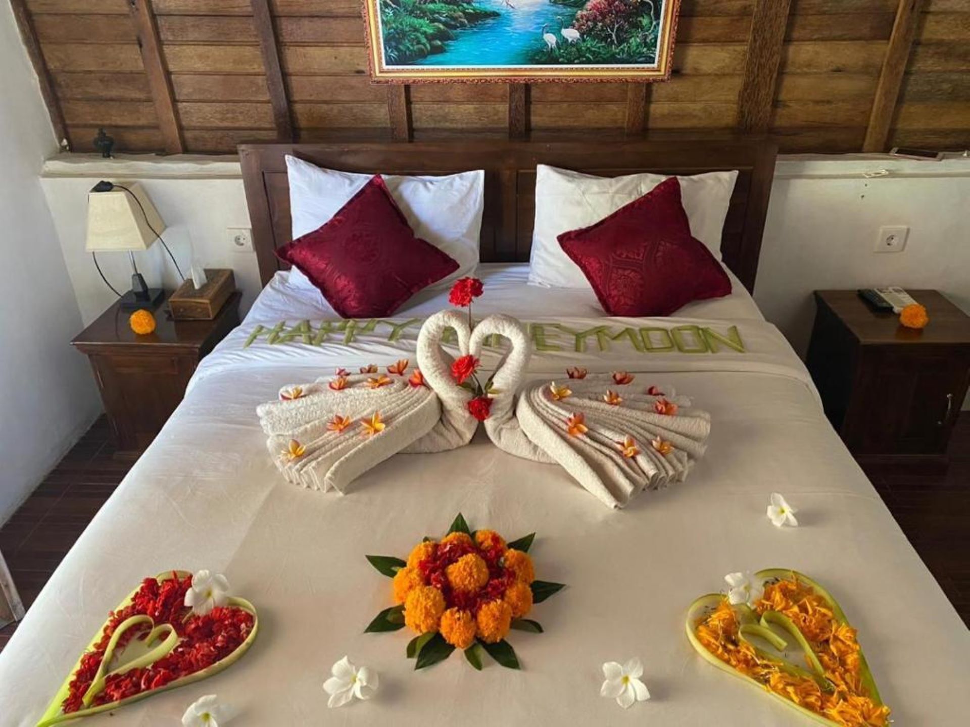 Habitación de hotel con una cama bien hecha, dos almohadas rojas, una mesita de noche con lámpara y un cuadro en la pared.
