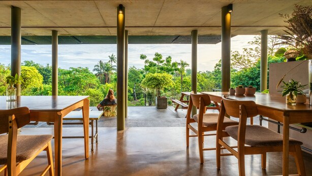 Un espace de restauration extérieur abrité, doté de tables et de chaises en bois, offrant une vue sur une végétation luxuriante et surmonté d’une structure en béton.