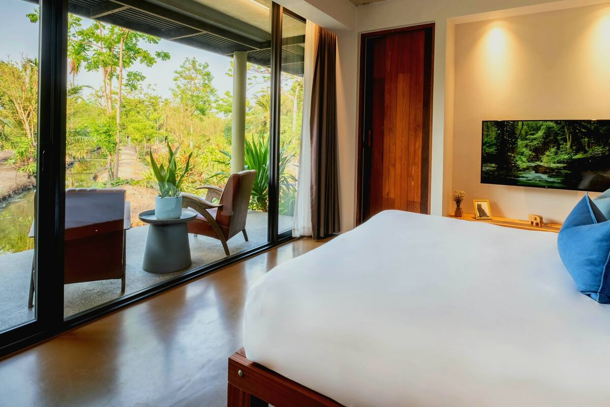A modern bedroom with a large bed, a TV mounted on the wall, and a view of a garden through floor-to-ceiling windows.