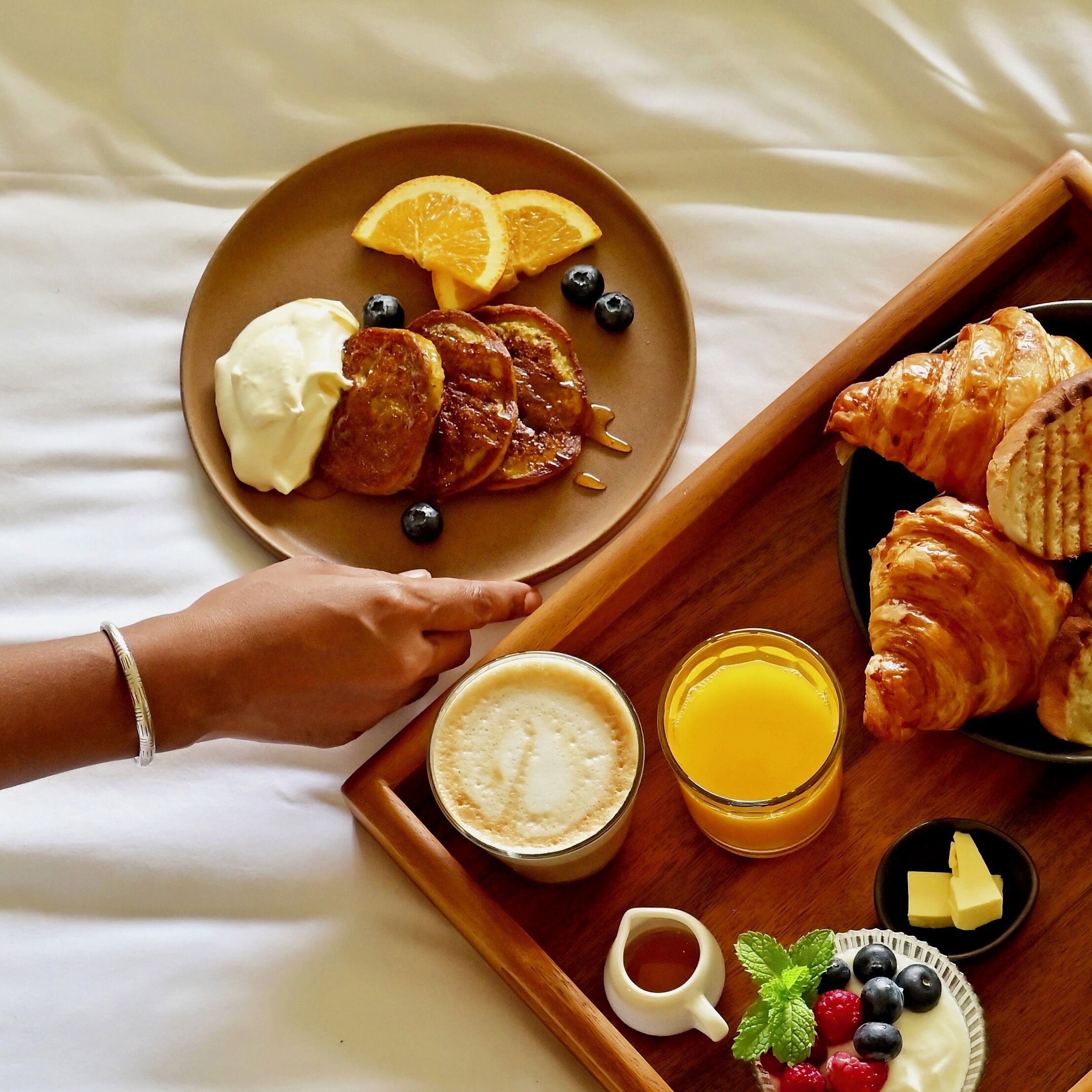 Bandeja de desayuno con tortitas, rodajas de naranja, arándanos, croissants, un vaso de zumo de naranja, una taza de café y un recipiente pequeño de mantequilla.