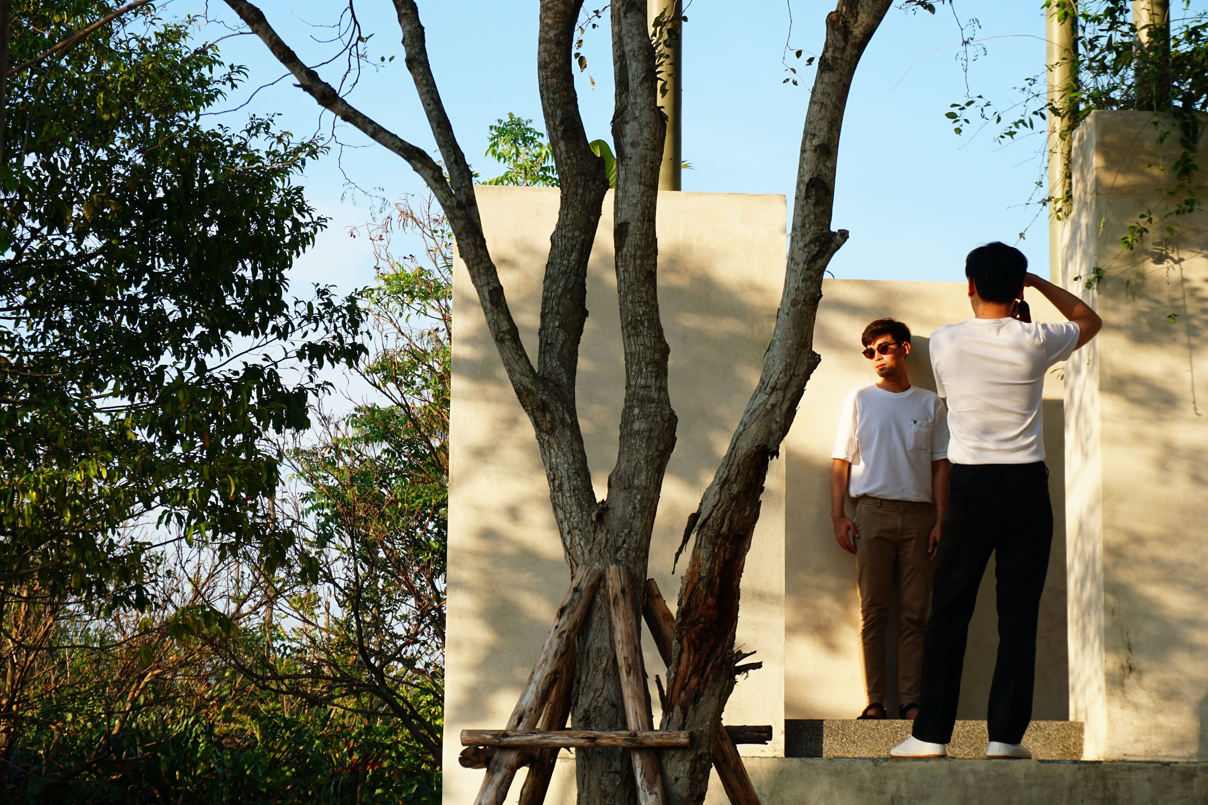 Dos personas de pie cerca de un árbol y una pared.