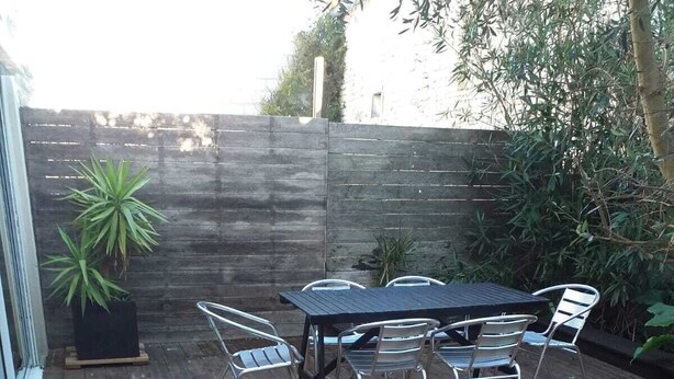 Un patio al aire libre con terraza de madera, una mesa redonda con sillas y plantas en macetas.