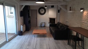 Una sala de estar moderna con piso de madera, un sofá de cuero negro, una mesa de centro de madera y un televisor. Hay una chimenea, un reloj y un área de comedor con sillas.