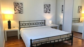 Un dormitorio con cama de estructura metálica, ropa de cama blanca, dos mesitas de noche con lámparas y obras de arte blanco y negro en las paredes.