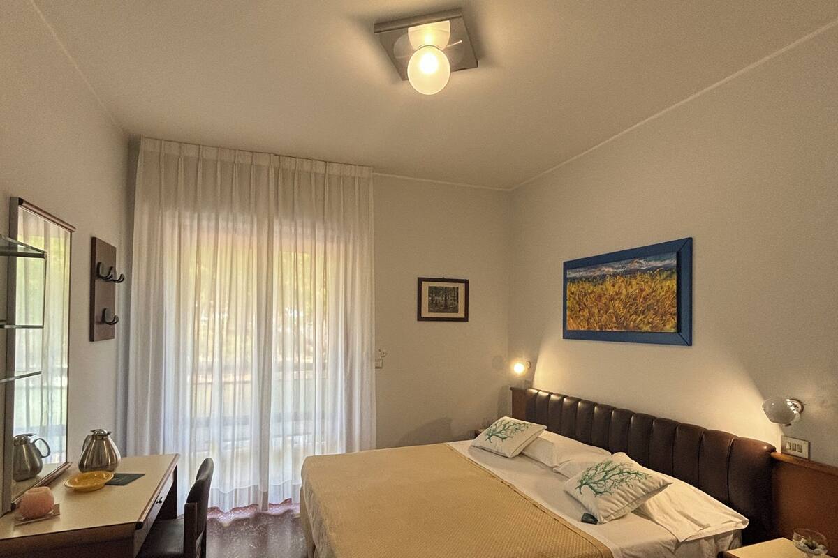 A hotel room with a bed, a desk, a chair, a window with curtains, a ceiling fan, and a painting on the wall.
