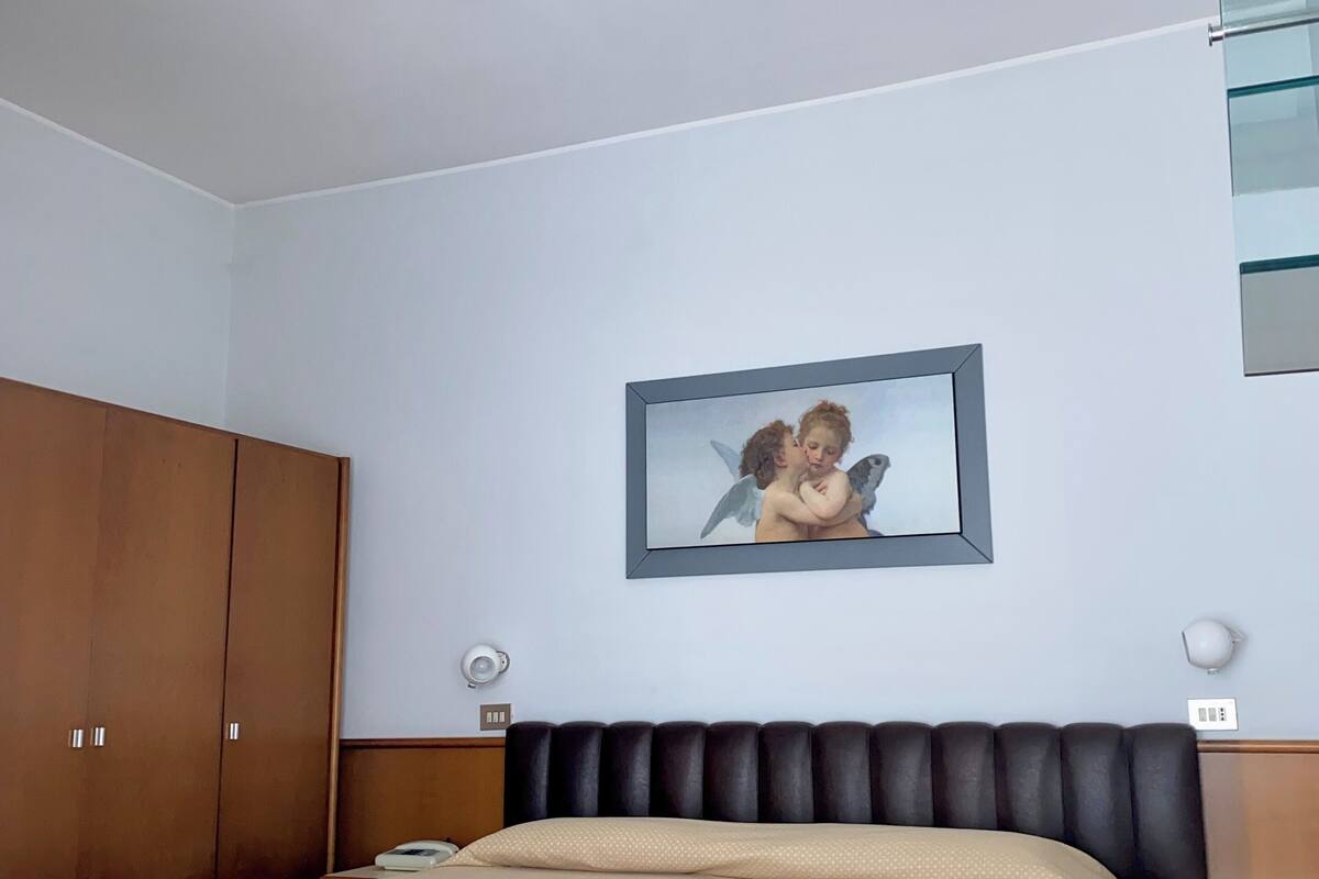 A hotel room with a bed, a wooden wardrobe, a framed picture on the wall, and a small table with a yellow bowl.