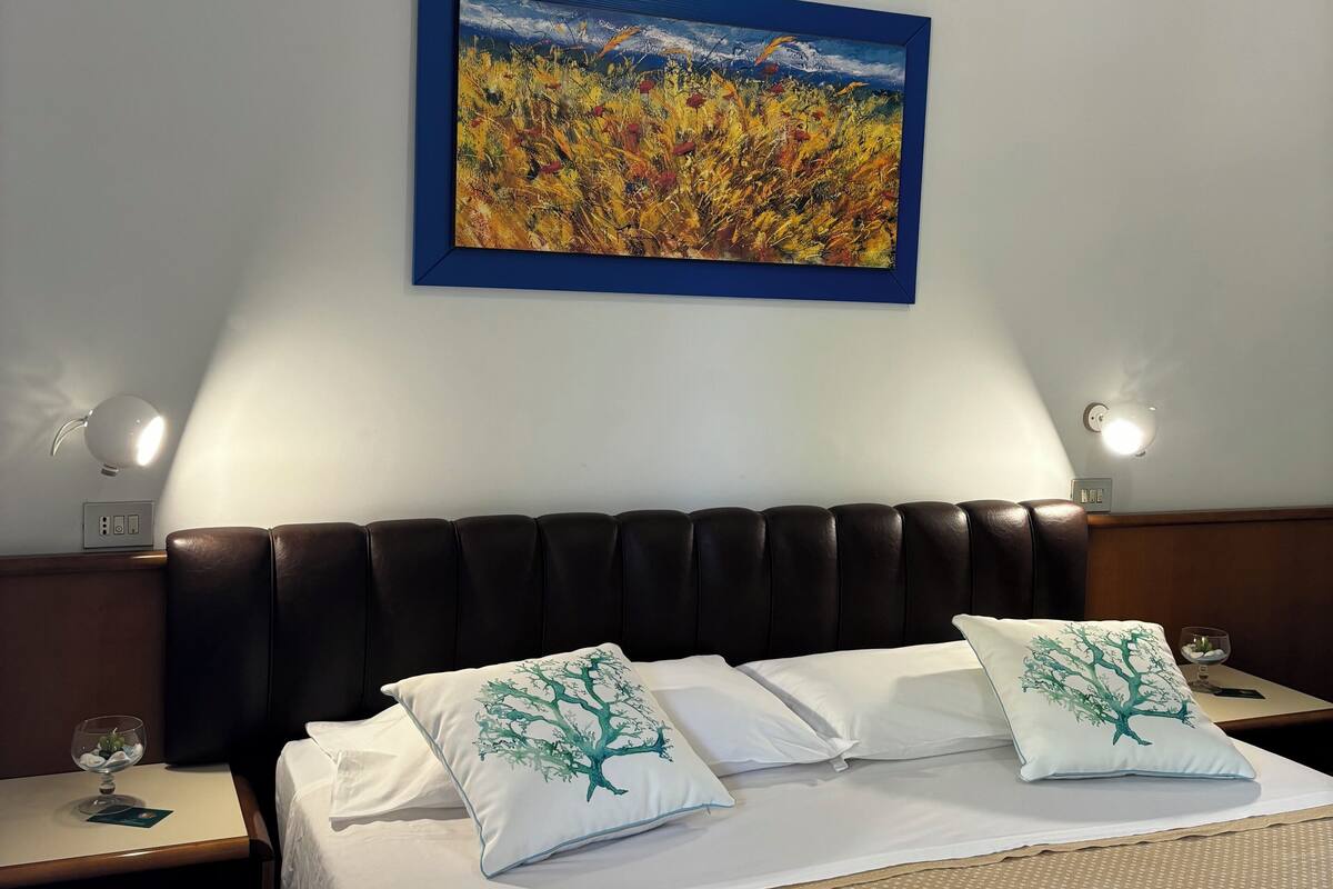 A hotel room with a bed, two pillows with tree designs, a nightstand with a glass and a book, a wall-mounted painting of a field, and a wall-mounted light.