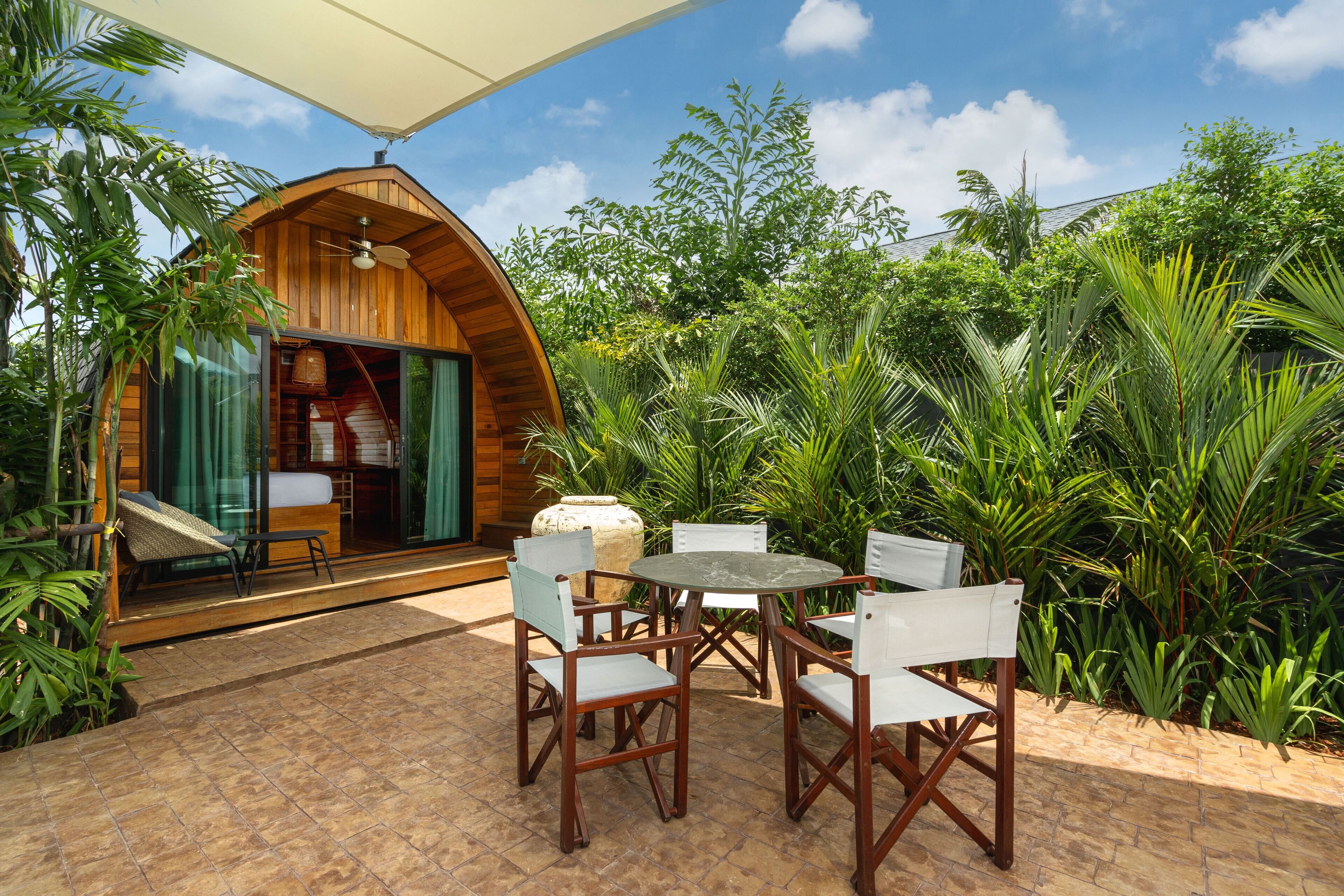 A wooden cabin with a glass door, a dining table with chairs, and lush greenery around.