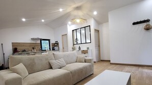 Ein geräumiges Wohnzimmer mit einer großen beigefarbenen Couch, einem weißen Couchtisch und einem gewebten Pouf. Im Hintergrund ist eine Küchenzeile mit modernen Geräten und einer Holzrückwand zu sehen.