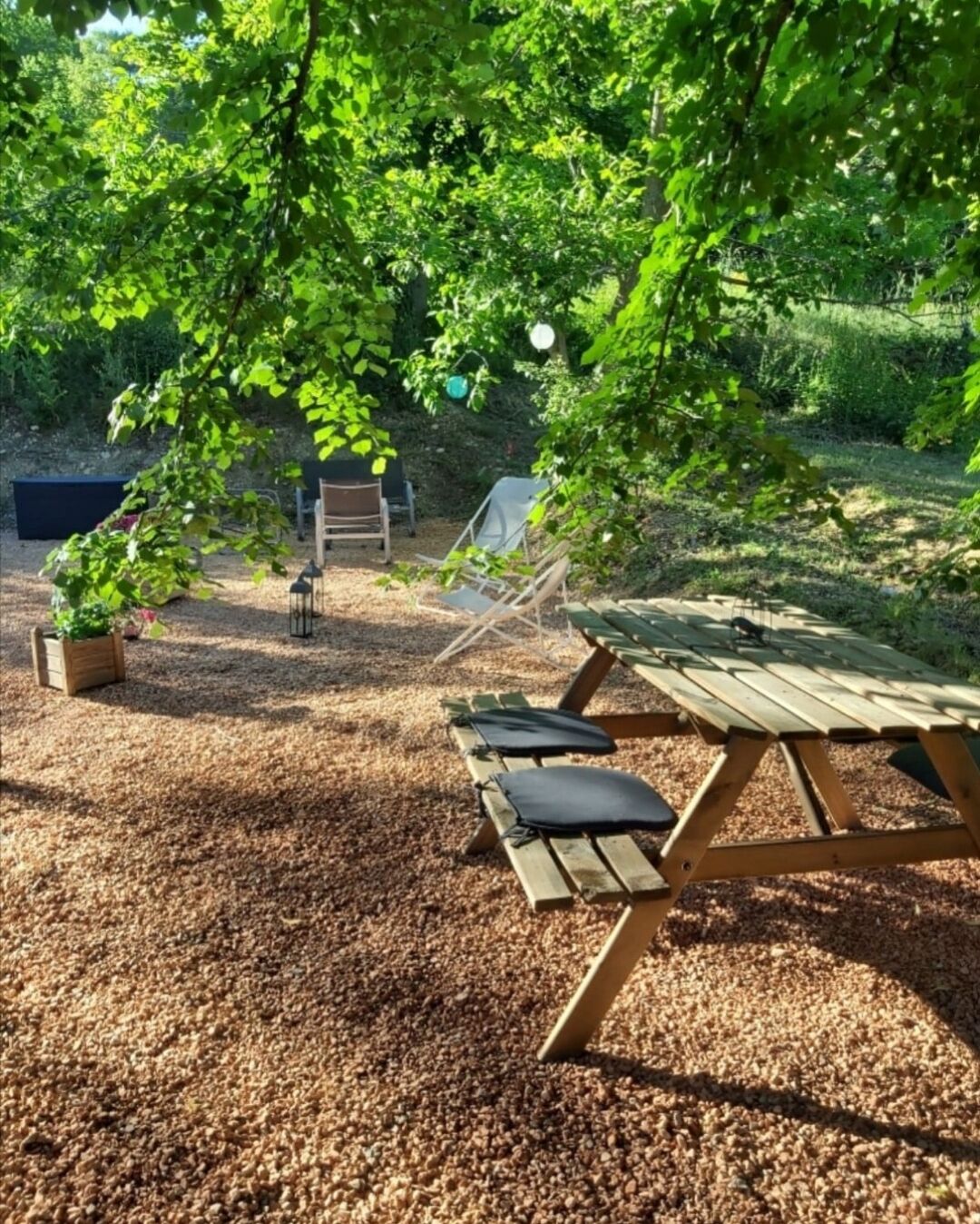 Une table de pique-nique en bois, avec des bancs, entourée d’une zone recouverte de gravier et de verdure.