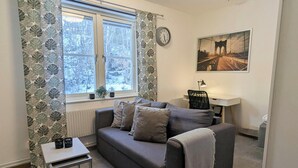 Un salon chaleureux avec un canapé gris, une petite table, une lampe, une horloge et une fenêtre donnant sur des arbres.