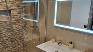 Un baño moderno con ducha envuelta en vidrio, un espejo grande con iluminación integrada, un tocador blanco con gabinete de madera y una pared con diseño de piedra.