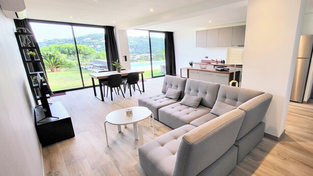 Una sala de estar moderna con un sofá seccional gris, una mesita blanca y un comedor con una mesa negra y sillas. Hay una zona de cocina con armarios y un refrigerador. Amplias puertas corredizas de vidrio dan a un balcón con vistas a zonas verdes y una piscina.
