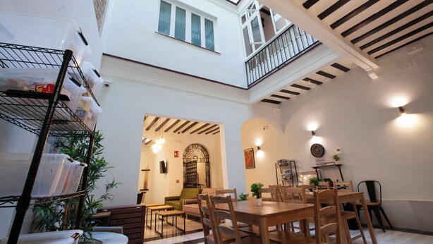 A spacious dining area with wooden tables and chairs, a staircase leading to an upper level, and a balcony with a railing. - Hostel Ibarra by Bossh! Hotels (Seville)