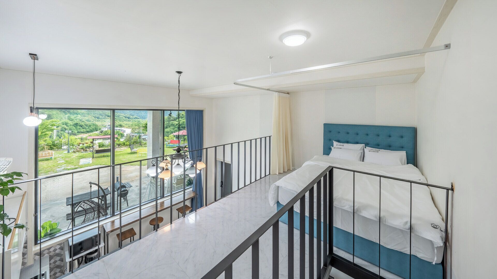 A hotel room with a balcony, a bed with a blue headboard, and a view of a green landscape.