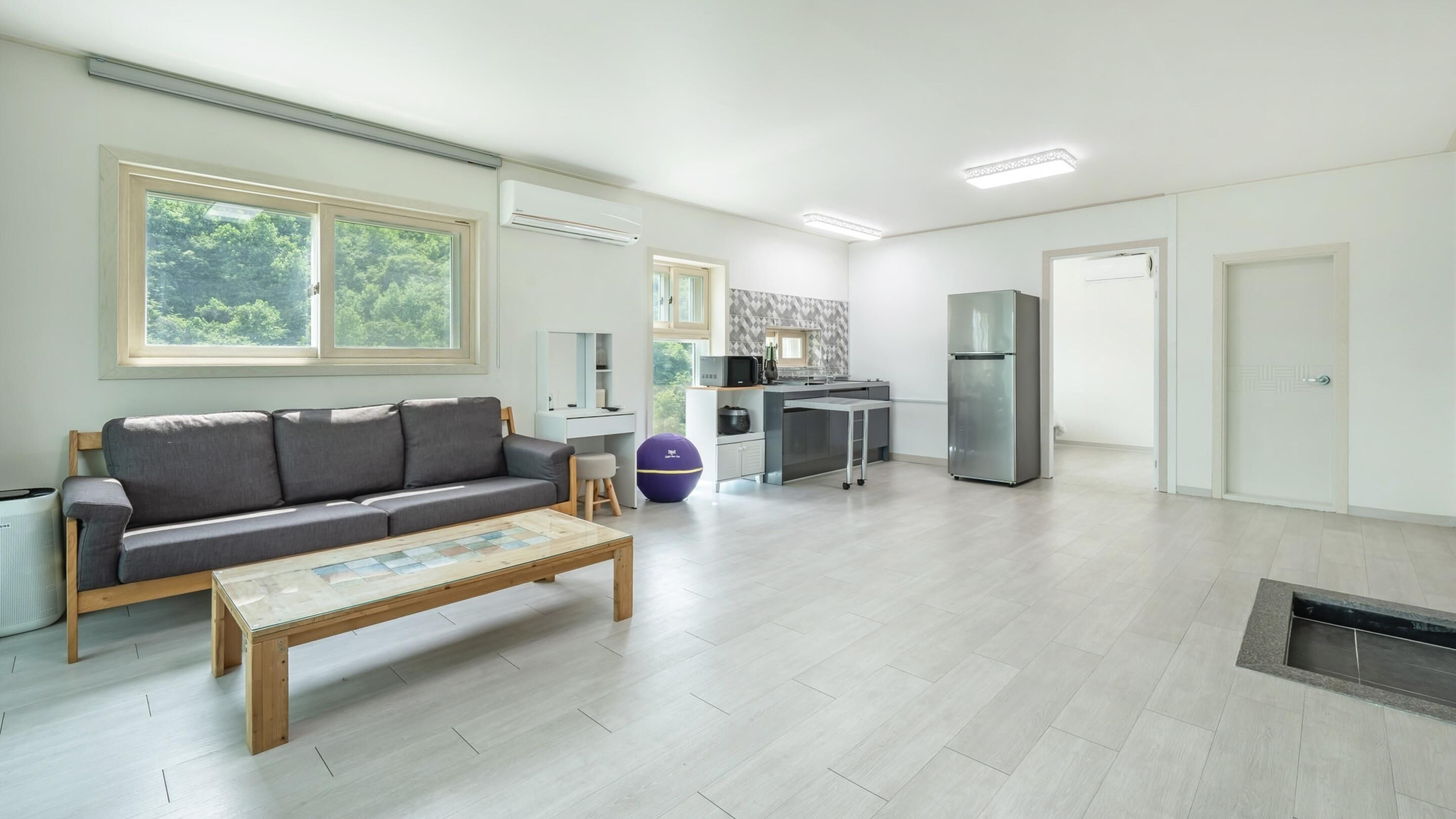 Amplia sala de estar con un sofá gris, una mesa de centro de madera y una cocina con refrigerador y microondas.