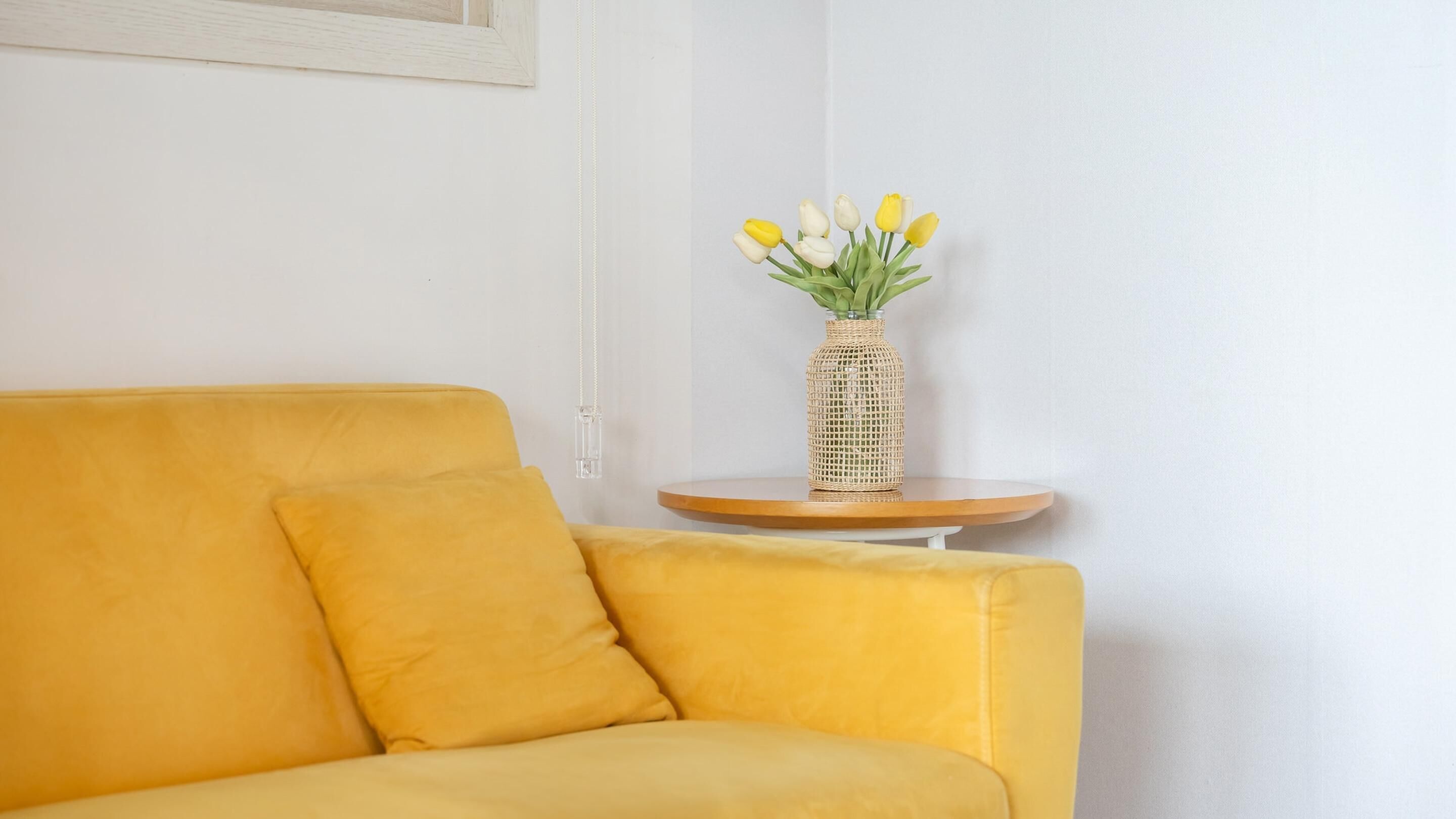 A yellow sofa with a cushion, a small round wooden table with a woven vase of yellow and white tulips, and a plain white wall.