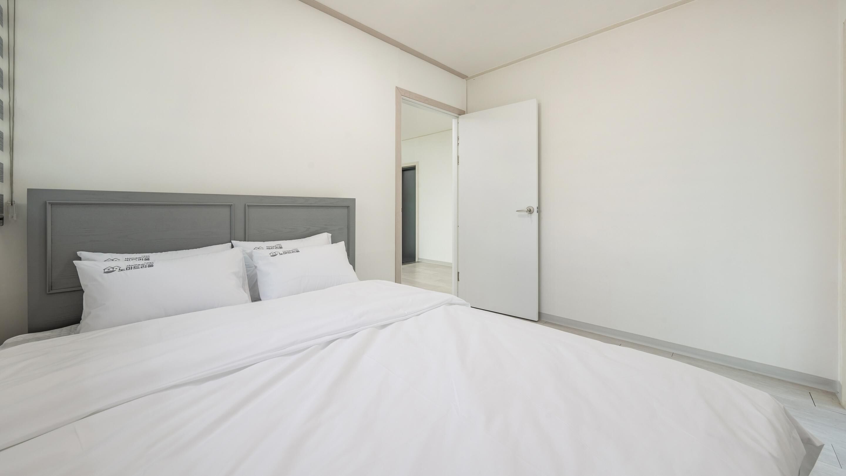 A modern, minimalist bedroom with a grey headboard, white bedding, and a white door leading to a hallway.