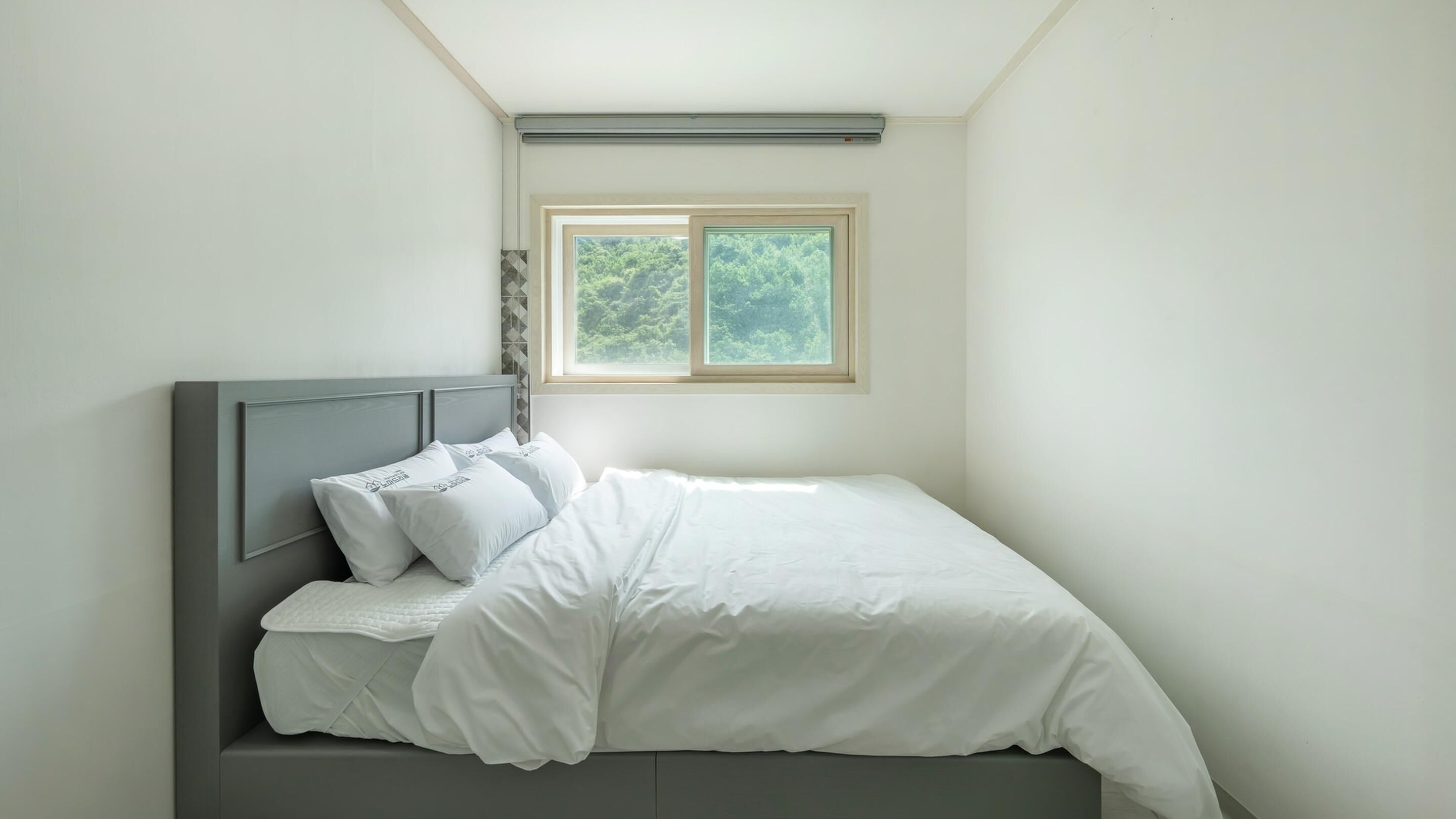 A neatly made bed with white bedding and pillows, a grey headboard, and a window with a view of trees.