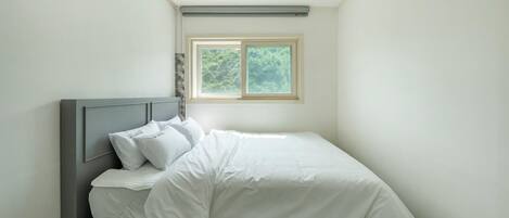 Una cama bien tendida con ropa de cama y almohadas blancas, un cabecero gris y una ventana con vistas a árboles.