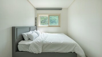 Una cama bien tendida con ropa de cama y almohadas blancas, un cabecero gris y una ventana con vistas a árboles.