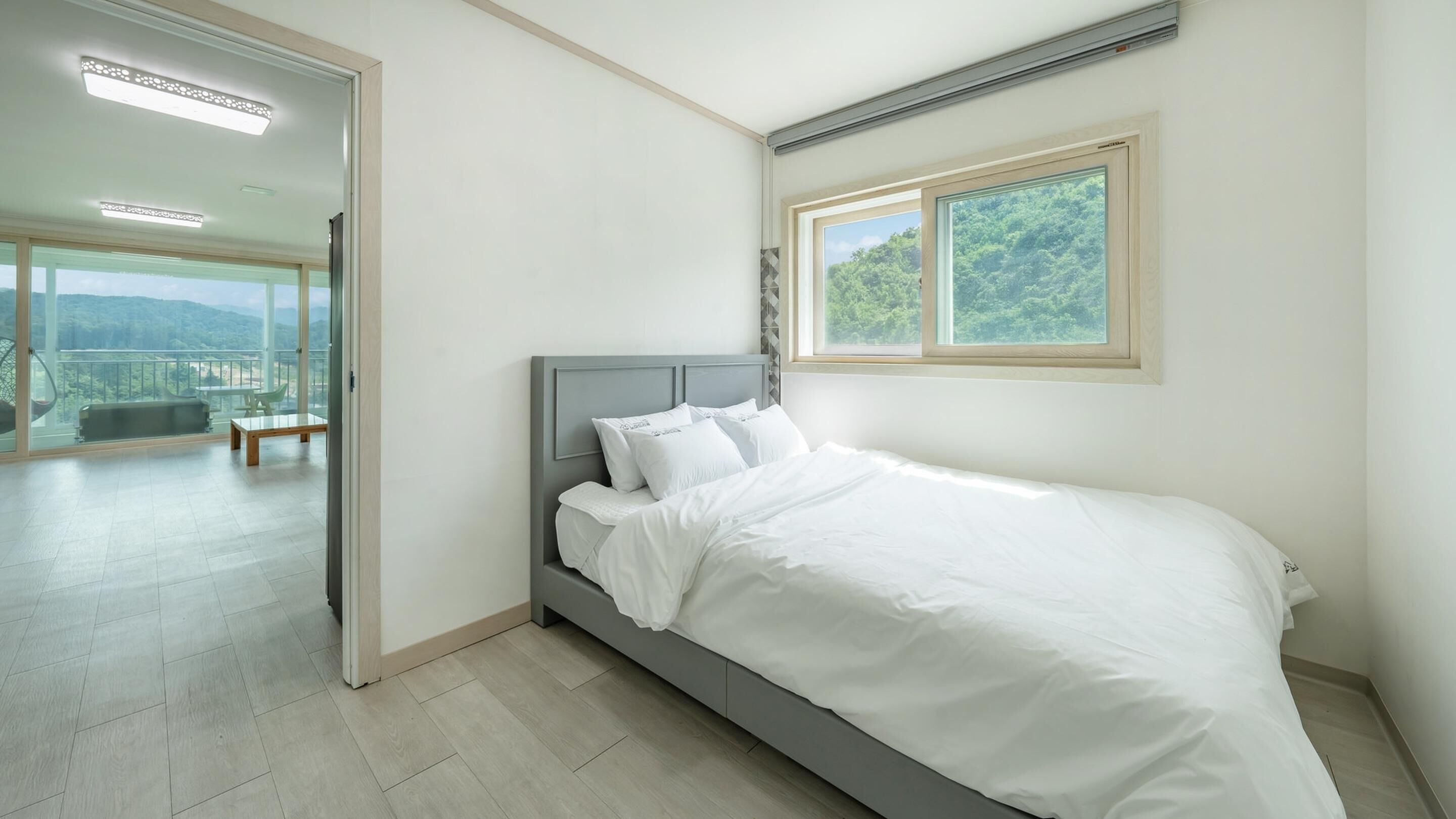 A bedroom with a bed, a window with a view of greenery, and a balcony with a table and chairs.