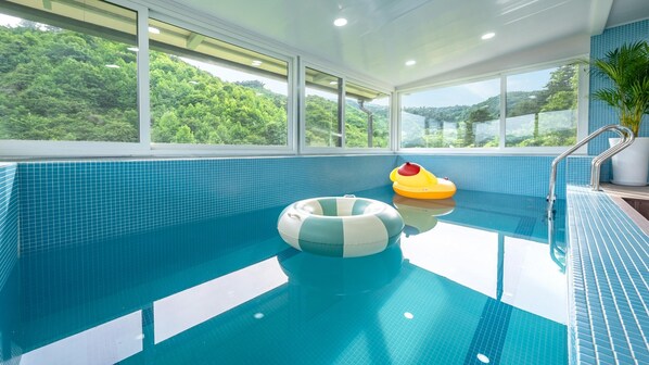 Piscina interior con baldosas azules brillantes, dos flotadores inflables y vistas a una vegetación exuberante a través de amplios ventanales.