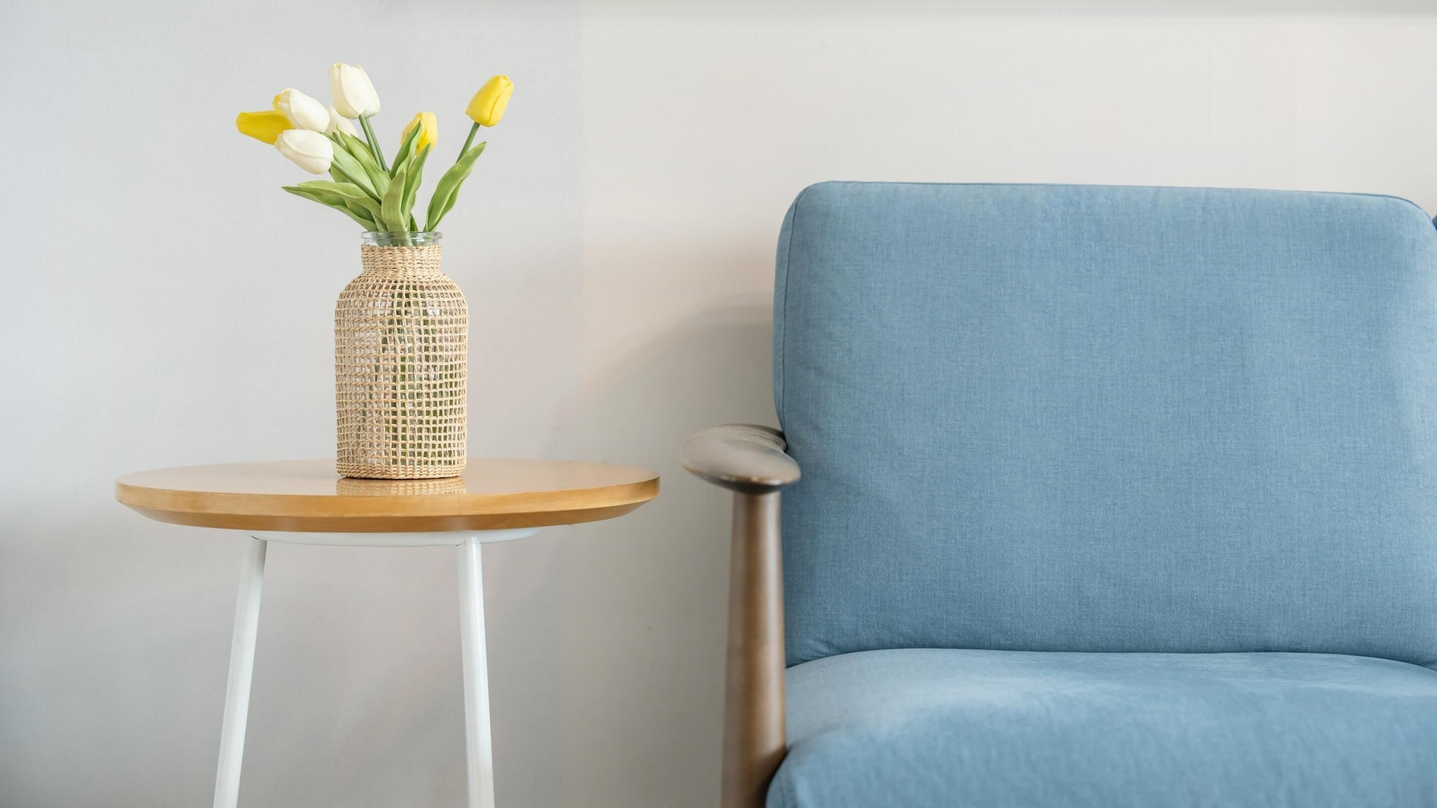 A round wooden table with a woven vase containing yellow and white tulips, positioned next to a blue upholstered chair.