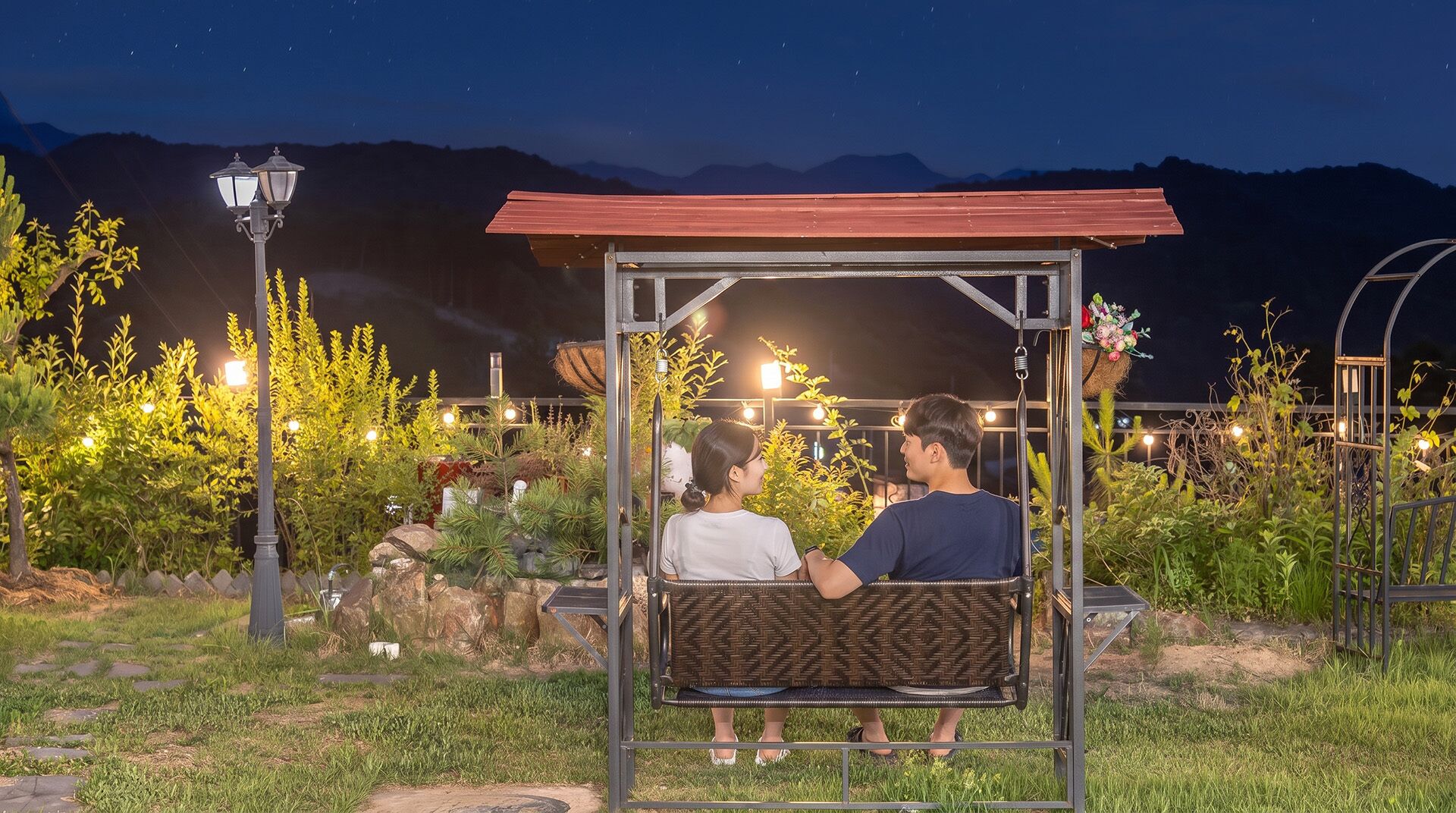 A couple on a swing in a garden with a metal gazebo, surrounded by greenery and a mountain backdrop.