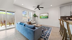 A modern living room with a blue sofa, a coffee table, and a TV. There is a dining area with wooden chairs and a table, and a kitchen with white cabinets and a microwave.
