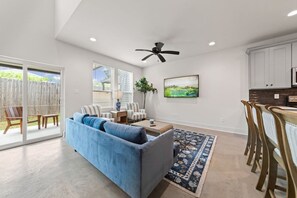 A modern living room with a blue sofa, a coffee table, and a TV. There is a dining area with wooden chairs and a table, and a kitchen with white cabinets and a microwave. - Cajun French House*2 King Beds*Fast Wi-Fi*Garage (Lafayette)