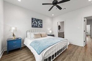 A bedroom with a white bed, a blue nightstand, a ceiling fan, a wall-mounted artwork, and a wooden floor. - Cajun French House*2 King Beds*Fast Wi-Fi*Garage (Lafayette)