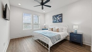 A bedroom with a white bed, a ceiling fan, two bedside tables with lamps, and a large window with blinds.