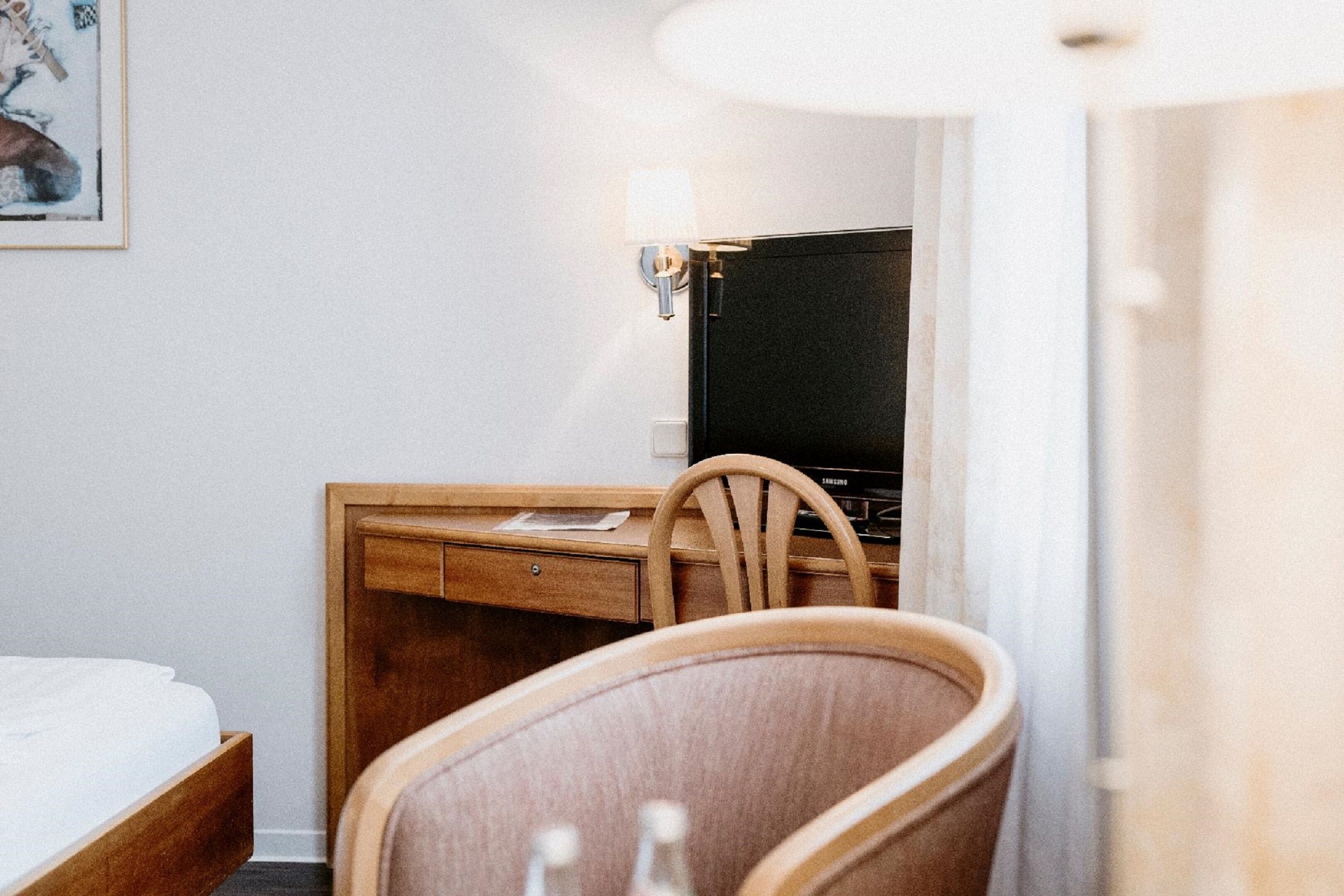 A hotel room with a bed, a wooden desk, a chair, a television, and a wall-mounted light fixture.