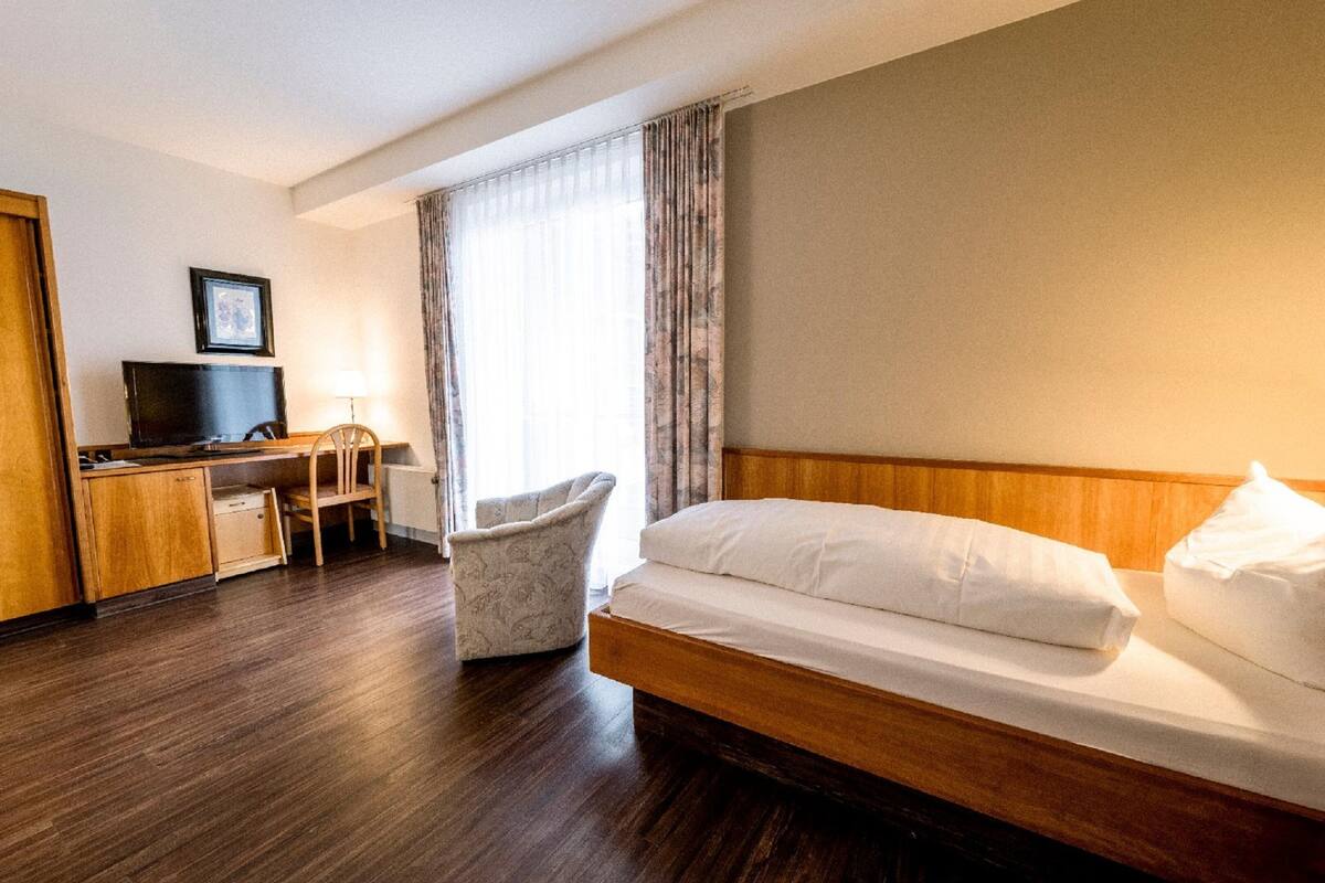 Ett hotellrum med en säng i trä, ett skrivbord med en stol, en tv och ett fönster med gardiner.