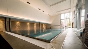 A modern indoor swimming pool with a glass wall, a textured wall on one side, and a clear view of the outdoors through large windows. - Kurhotel San Andreas (Bad Fuessing)